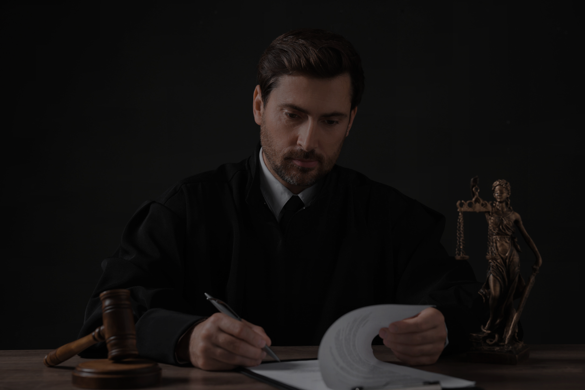 Judge with gavel writing in papers at wooden table against black background